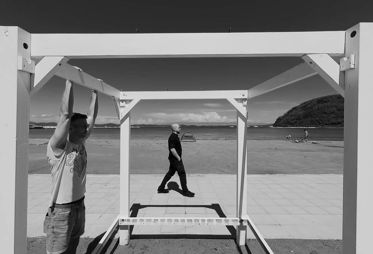 JM Kurobolja – No F*cks Given Black and white photo of a man hanging from a wooden structure by the beach with another man walking on the pavement behind him.