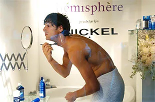 JM Public shower shave A young man with a towel around his waist shaving his face in a bathroom mirror, surrounded by grooming products.