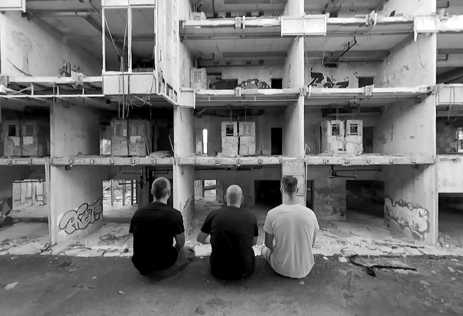 JM Re:Play Three men sit with their backs to the camera facing a large, abandoned, graffiti-covered concrete building interior.